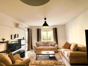A living room with a sofa, armchair, coffee table, TV, and a large window with curtains. - 2 bedrooms apartment in rehab city (New Cairo)