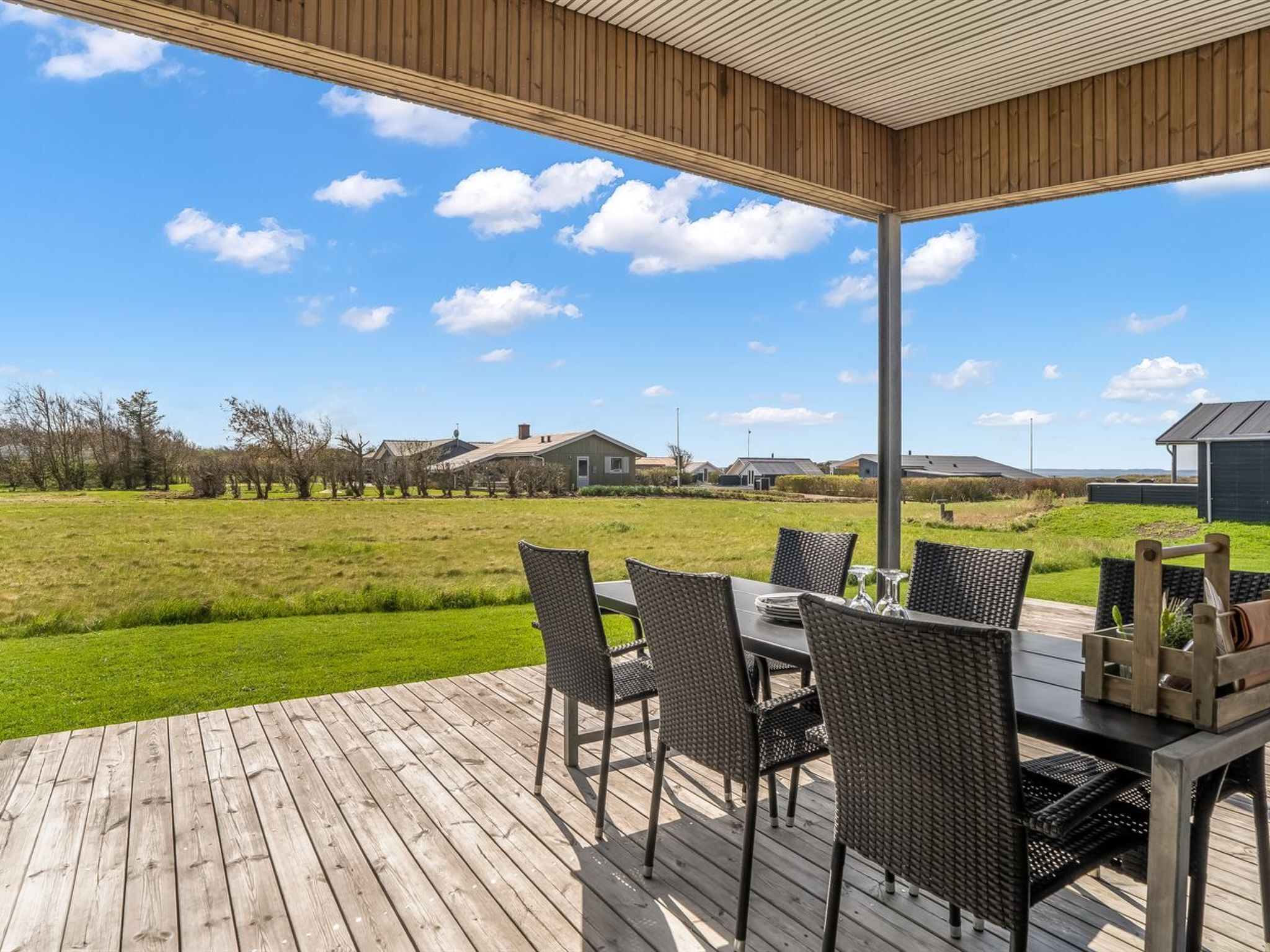 A covered outdoor dining area with a table and chairs, a view of a grassy field, and houses in the background.
