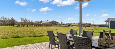A covered outdoor dining area with a table and chairs, a view of a grassy field, and houses in the background.