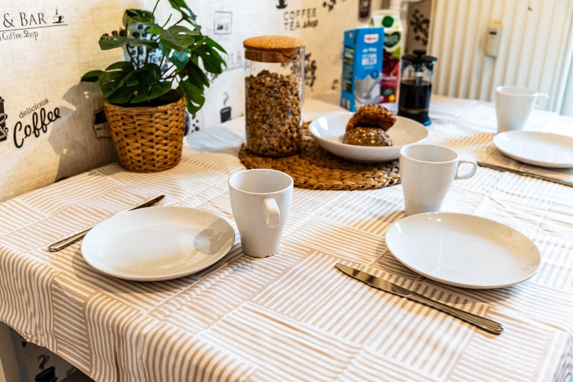 Ein Frühstücksarrangement mit Tellern, Tassen und einer Kaffeemühle auf einem Tisch mit gestreiftem Tischlauf.