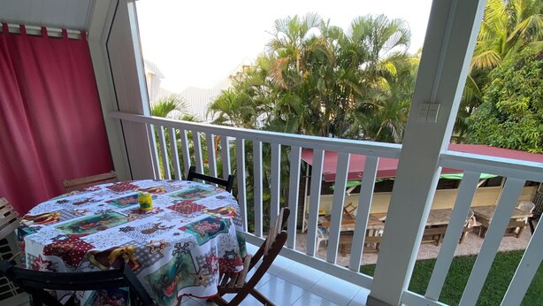 A balcony with a table and chairs, a red curtain, and a view of greenery and buildings. - Sainte-Anne | Pointe Marin beach 400m away! (Sainte-Anne)