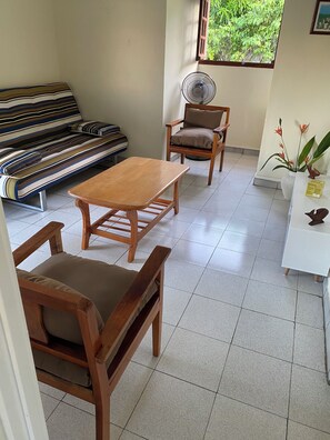 Ein Zimmer mit Holztisch und Stühlen, einer Couch, einem Standventilator und einem Fenster mit Blick auf Grünflächen.