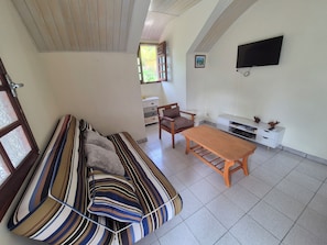 A living room with a striped sofa, a wooden table, a TV mounted on the wall, and a window with a view of greenery. - Sainte-Anne | Pointe Marin beach 400m away! (Sainte-Anne)