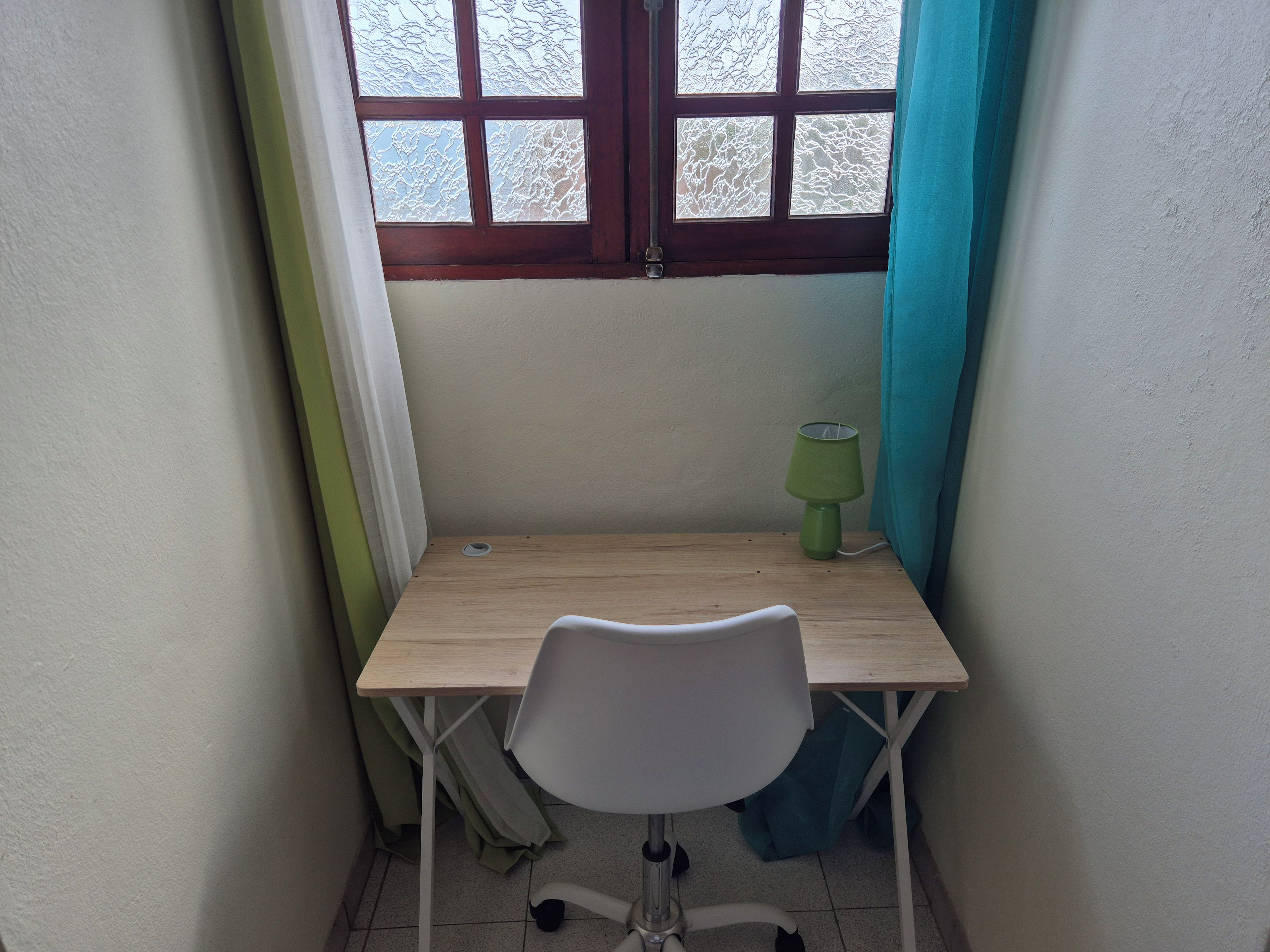 A corner room with a wooden desk, a white chair, a green desk lamp, and a window with frosted glass.