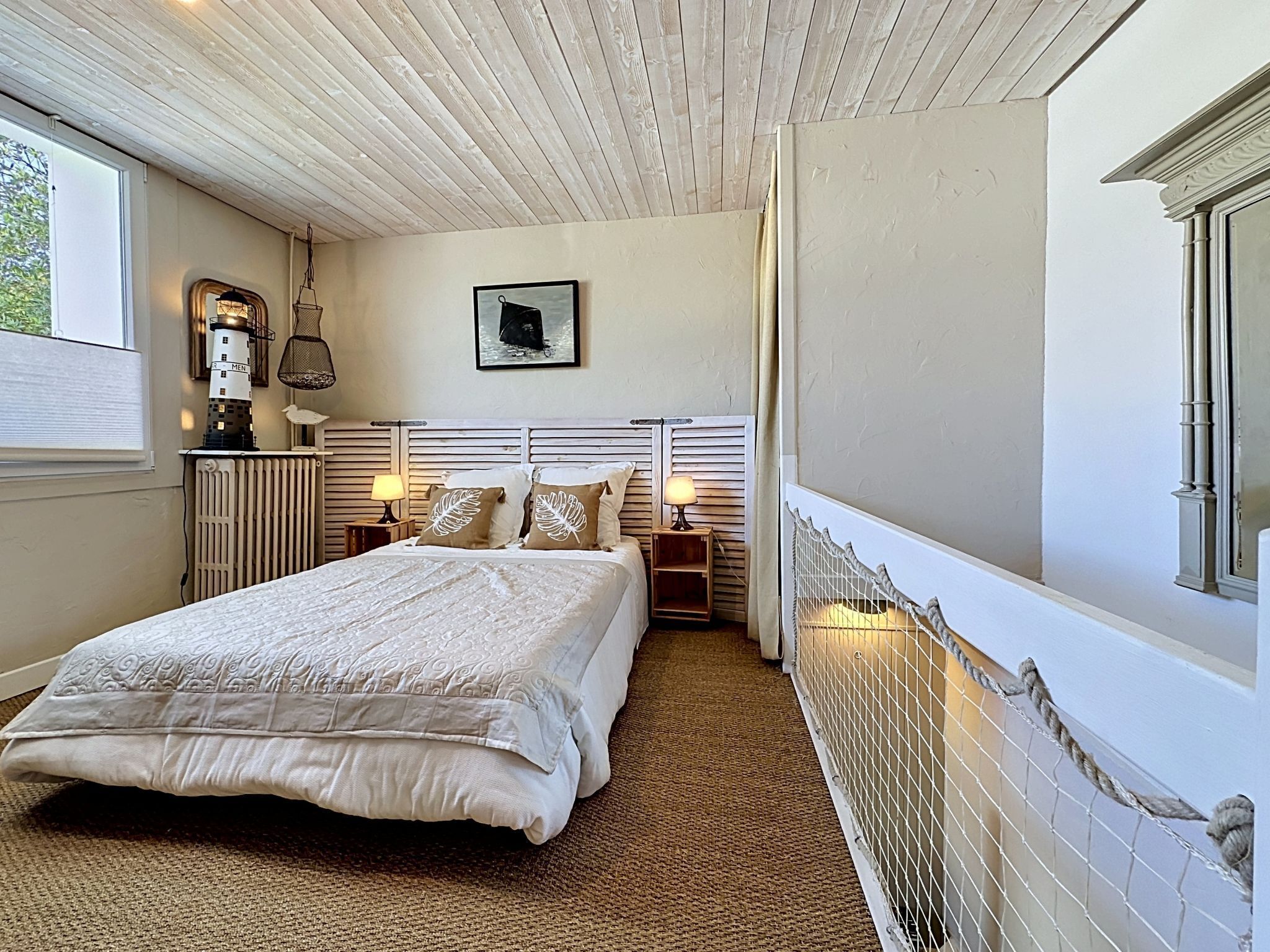 A bedroom with a large bed, a nightstand, a lamp, a framed picture on the wall, and a window with a view of a lighthouse.