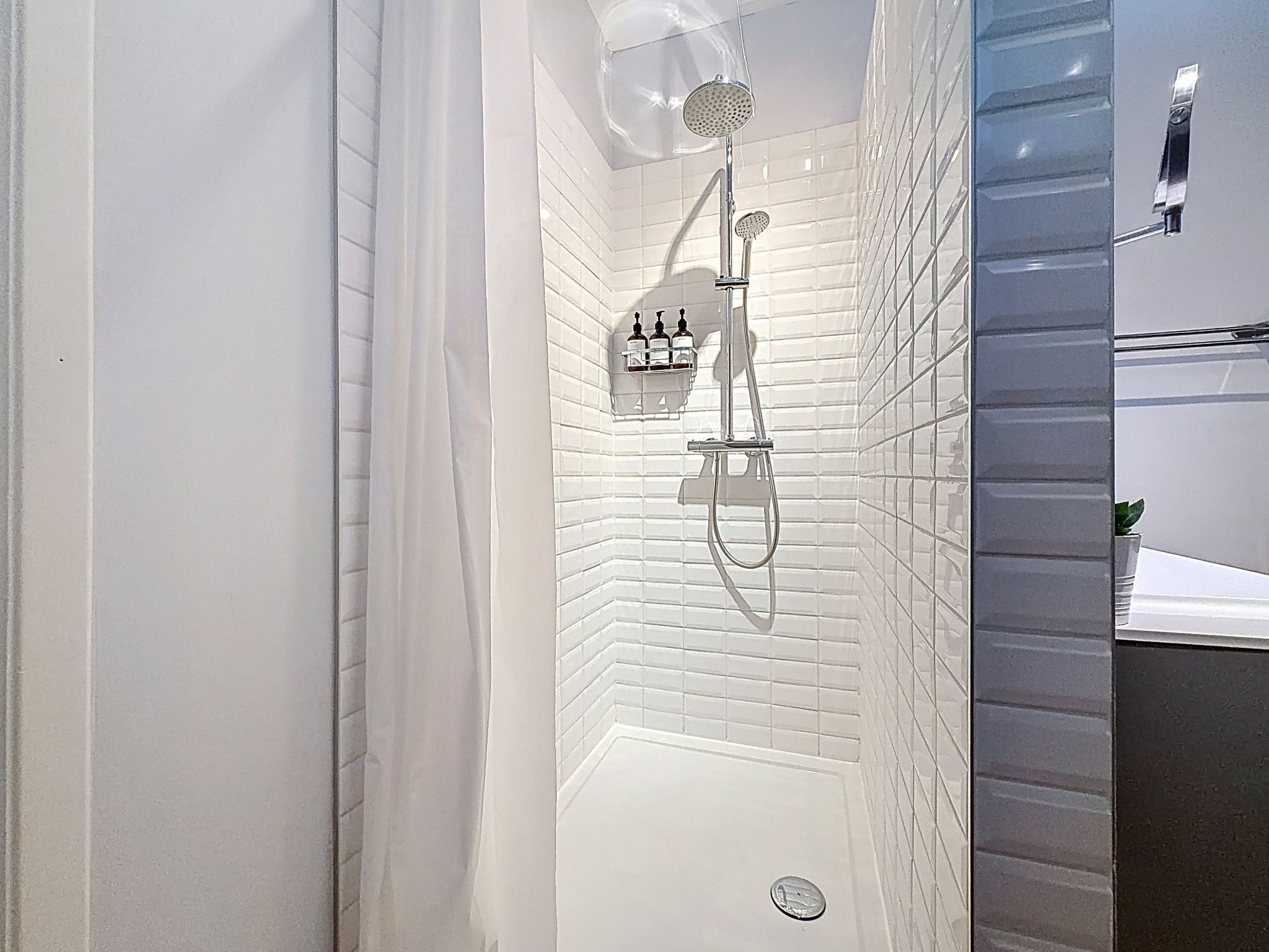 Une salle de bain moderne avec des murs recouverts de carreaux blancs, une douche équipée d’un pommeau de douche à main et d’un distributeur de savon encastré.
