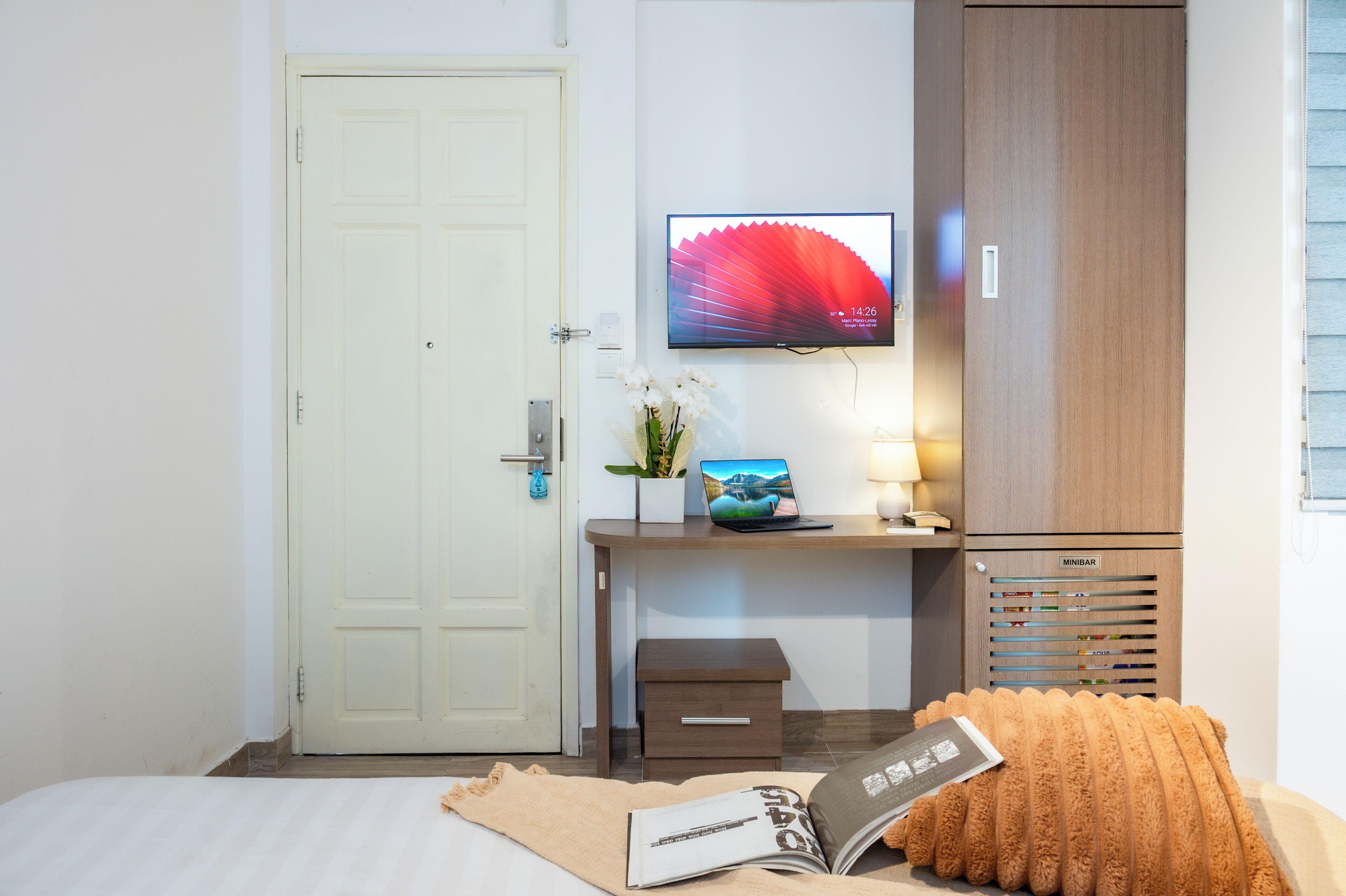 A hotel room with a bed, a desk with a laptop, a television, a Minibar, and a door.