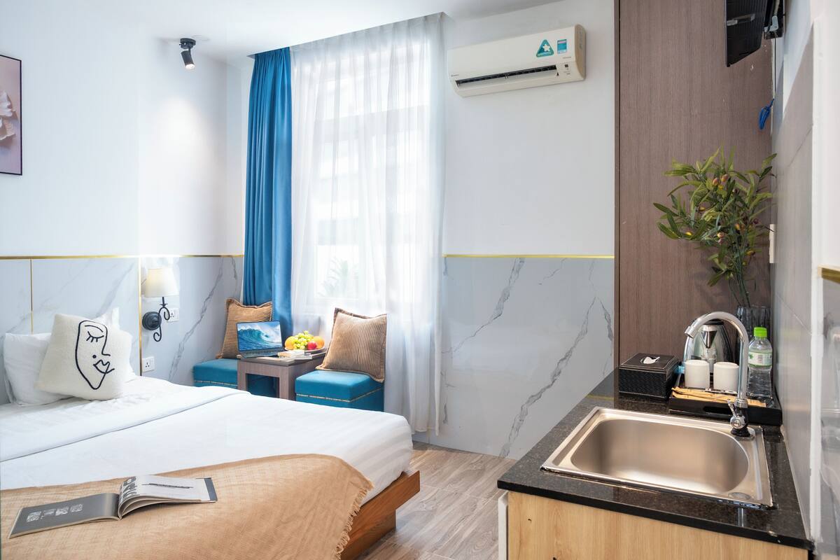 A hotel room with a bed, bedside table, lamp, air conditioning unit, and a kitchen area with a sink and countertop.