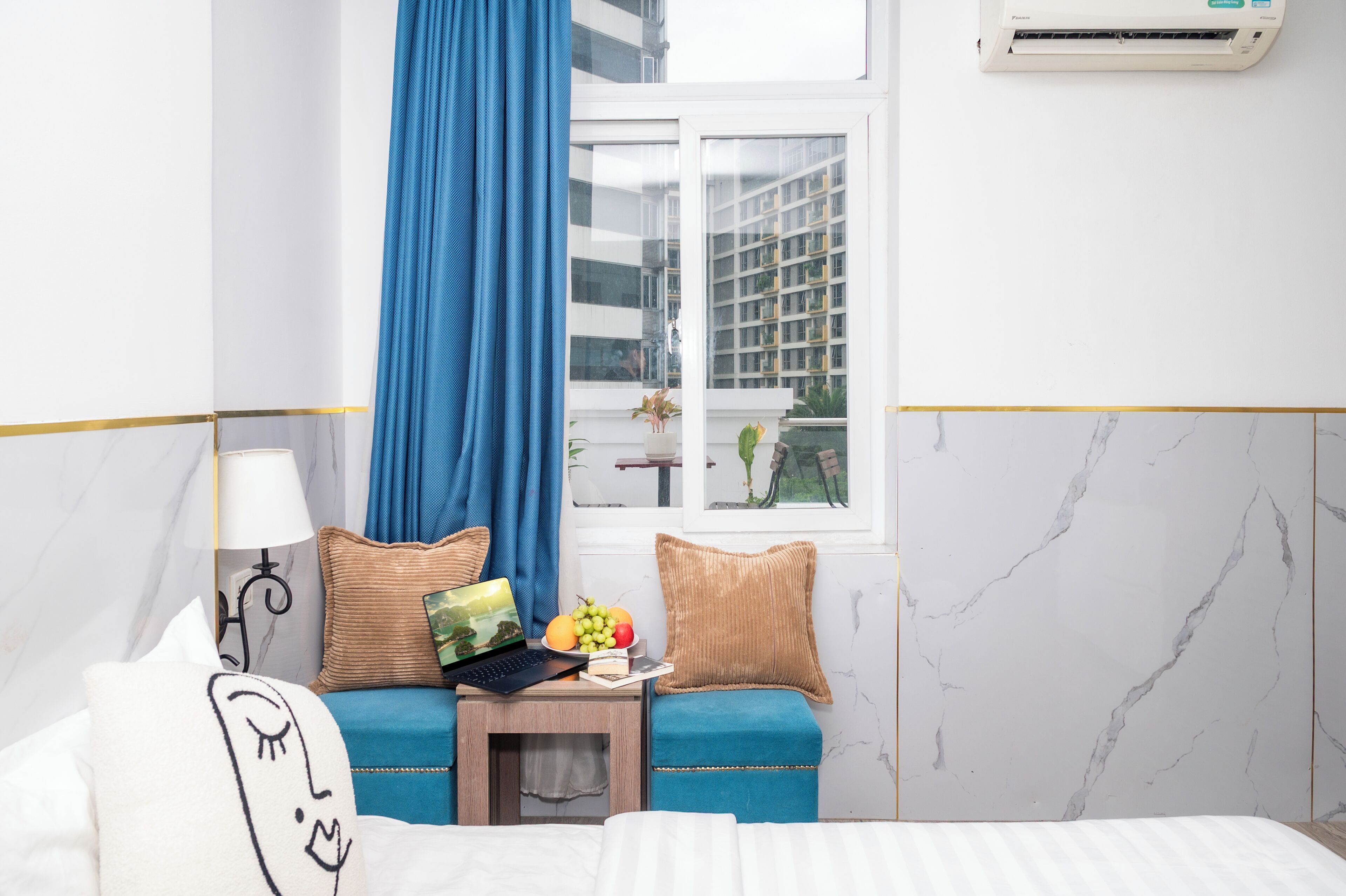 A hotel room with a bed, a desk with a laptop, a small table with fruit, a window with a view of buildings, and an air conditioner.