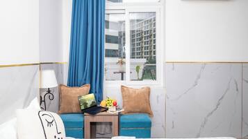 A hotel room with a bed, a desk with a laptop, a small table with fruit, a window with a view of buildings, and an air conditioner.