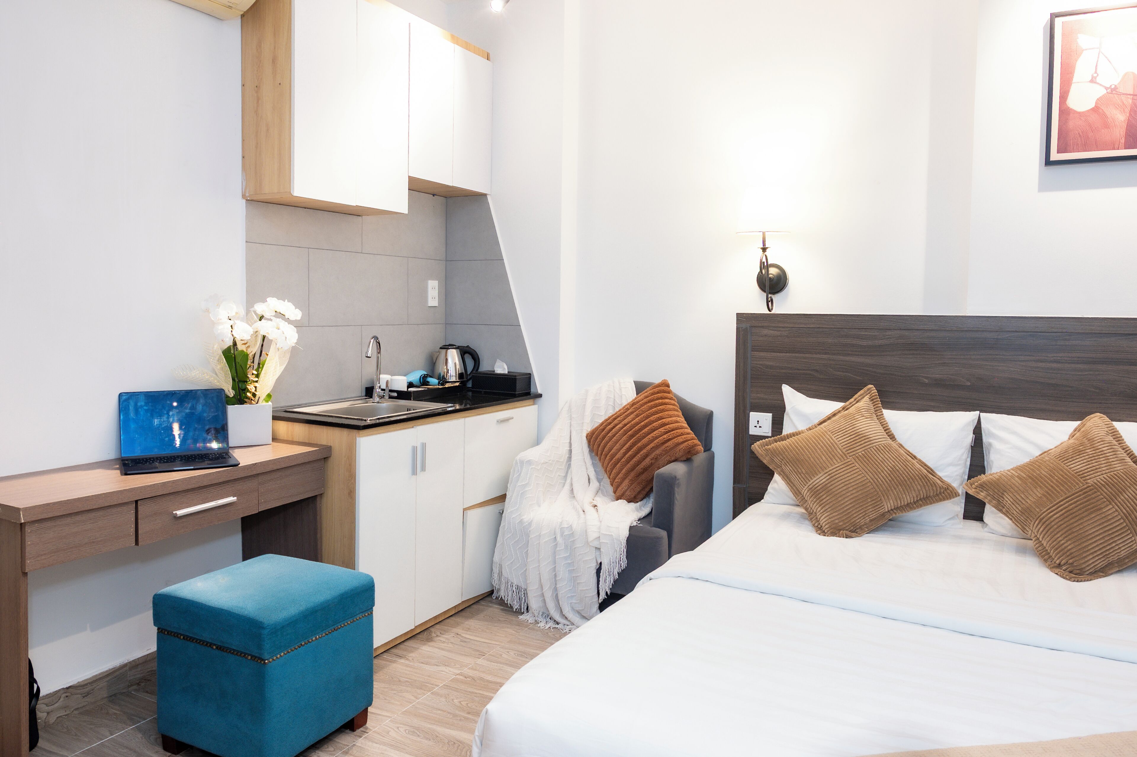 A modern hotel room with a kitchenette, a bed with brown pillows, a blue ottoman, and a wall-mounted lamp.
