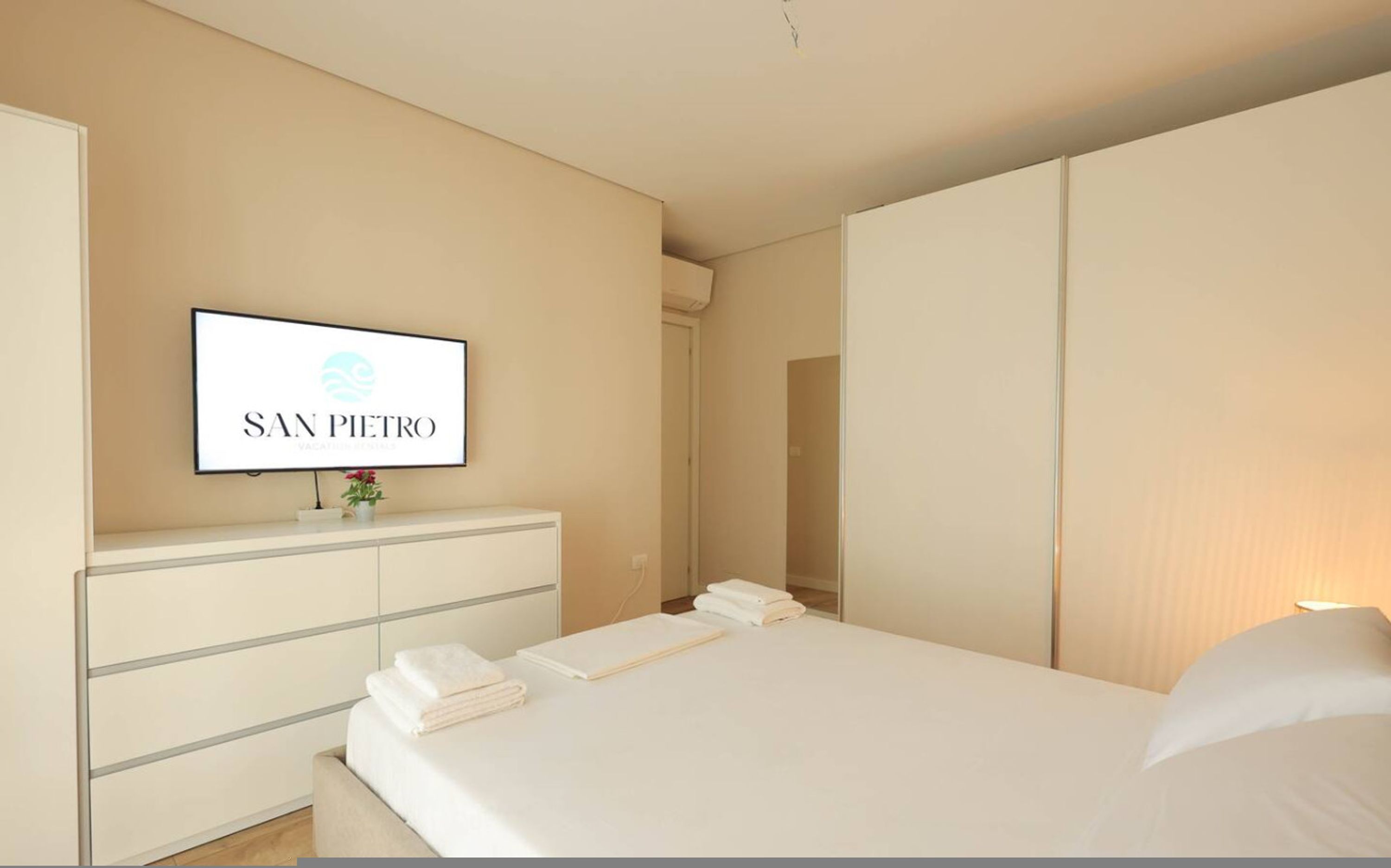A hotel room with a bed, a TV mounted on a wall, a wardrobe, and a bedside table with a vase of flowers.