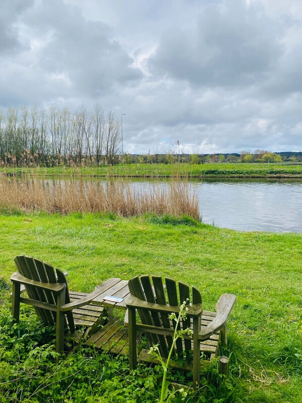 Two wooden Adirondack chairs facing a calm lake with a grassy bank and trees in the background.
