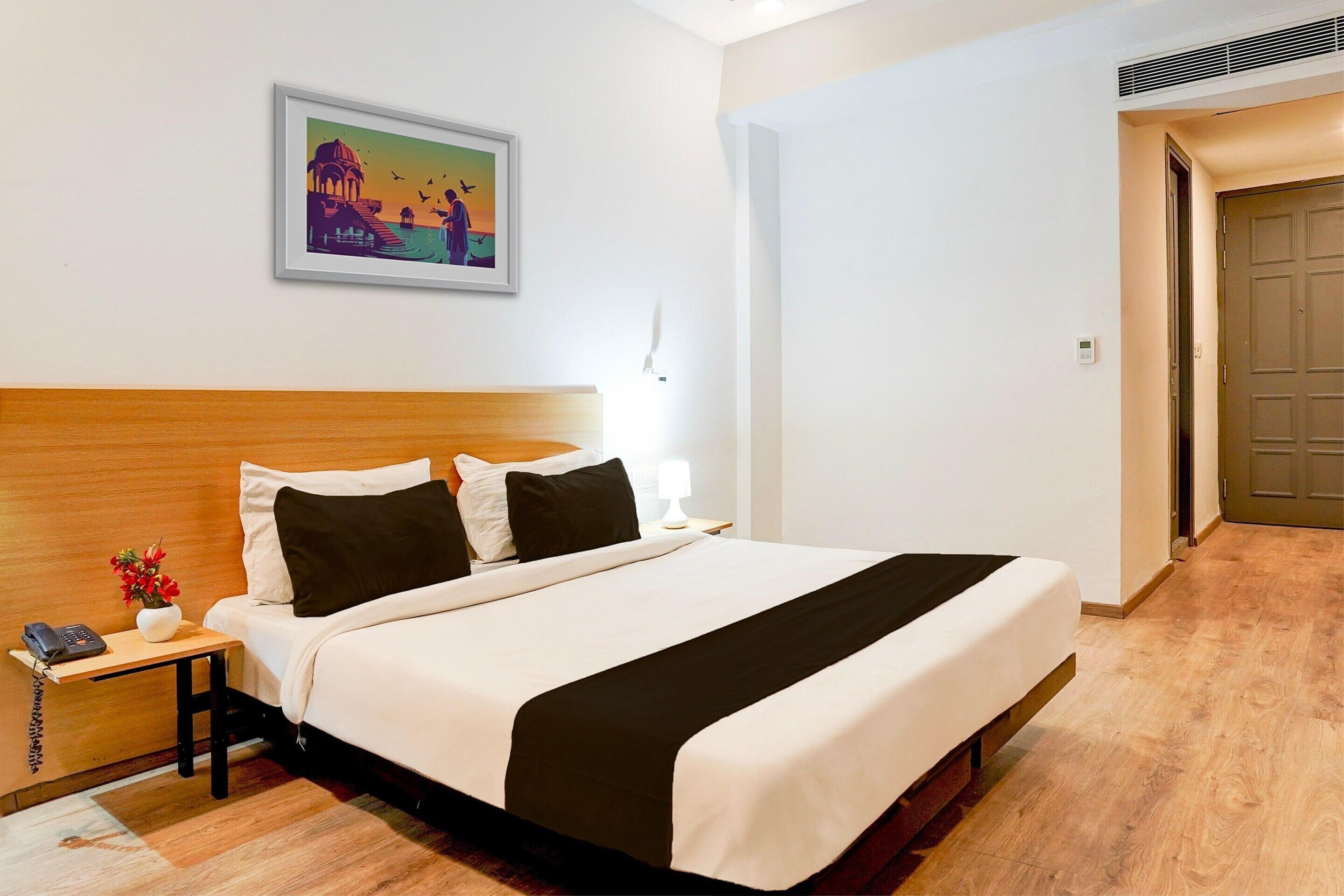 A hotel room with a bed, a nightstand with a phone, a framed picture on the wall, and a door to the right.