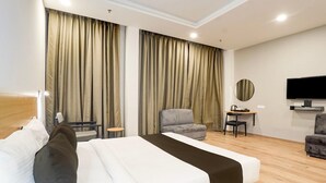 Hotellihuone, jossa on sänky, verhot, televisio, tuoli ja pieni pöytä.
