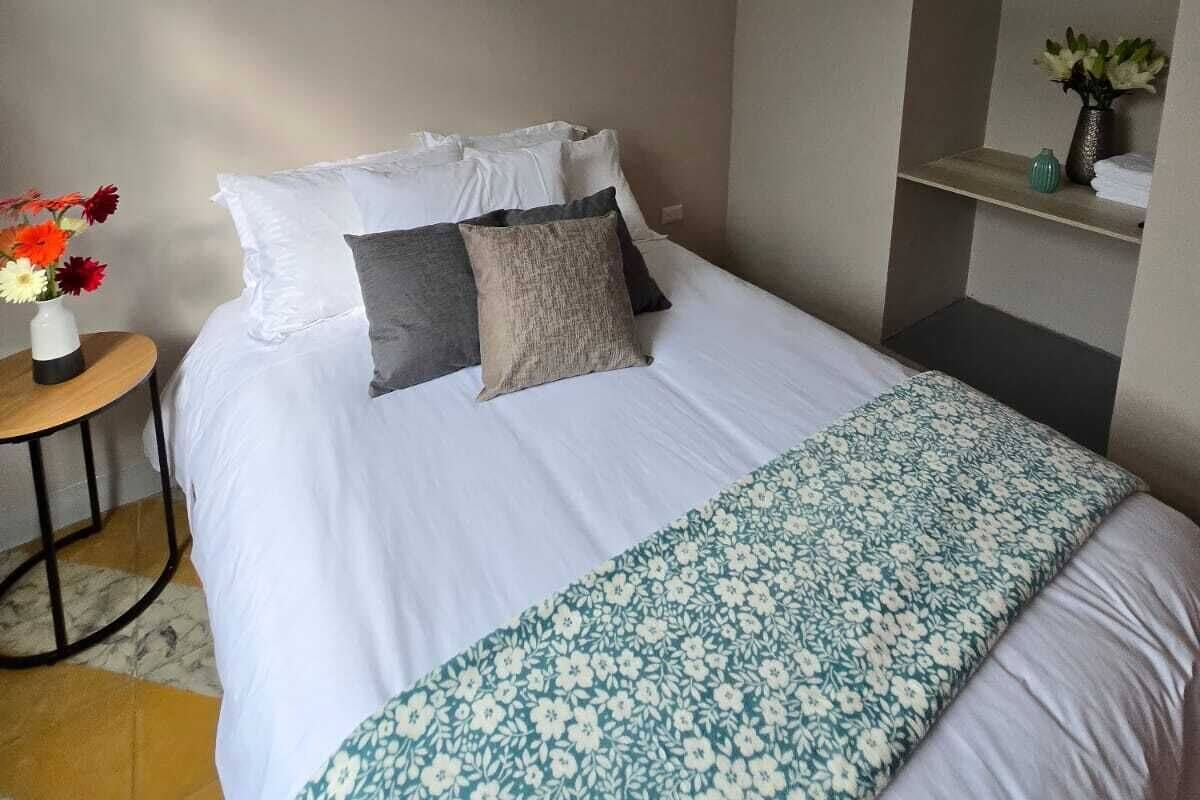 Una cama bien hecha con ropa de cama blanca y un plaid estampado floral, una mesita redonda con un jarrón de flores, y un espejo en la pared.