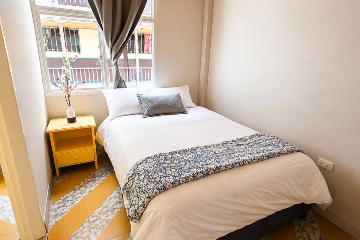 Un dormitorio con cama, mesita de noche, ventana con cortinas y vistas a edificios.
