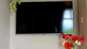 A flat-screen TV mounted on a wall, a white shelf with decorative items, a small white vase with flowers, and a wooden table.