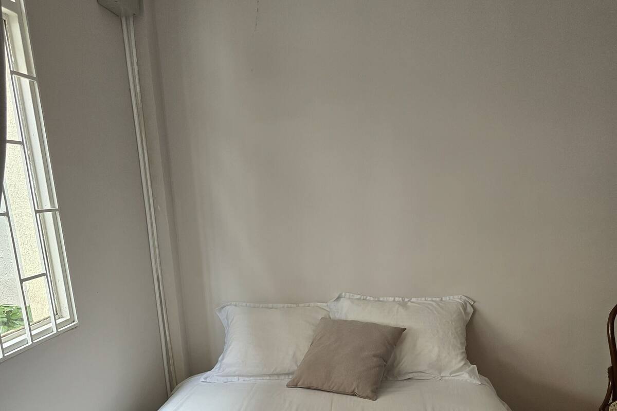 Una cama individual con ropa blanca y una manta gris, una ventana con marcos blancos, y una silla de madera.