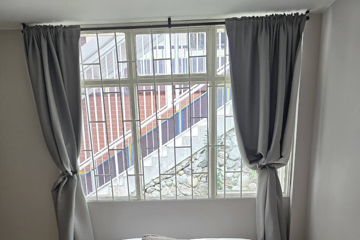Un dormitorio con cama, cortinas grises y ventana con vista a un edificio.