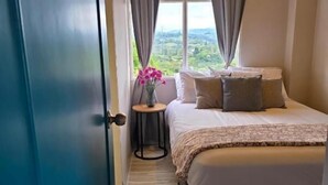 A bedroom with a bed, pillows, a small table with a vase of flowers, and a view of greenery outside.