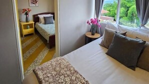 A hotel room with a wooden ceiling, a large bed with a floral bedspread, a small bedside table with a vase of flowers, and a view of greenery through a window.