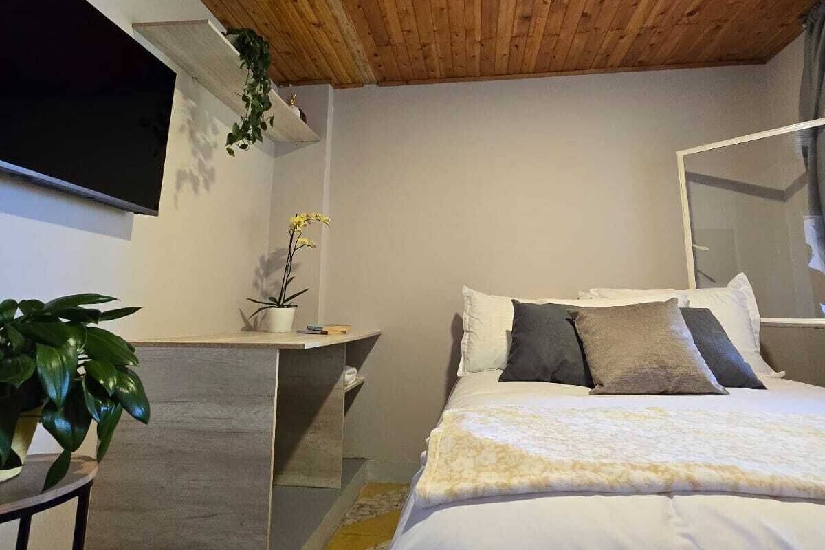 Un dormitorio con techo de madera, una cama con ropa de cama blanca, una mesita de noche con una planta y un televisor montado en la pared.