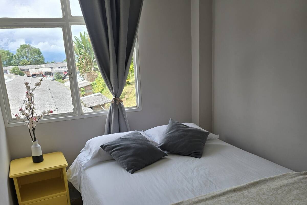 Un dormitorio con cama, mesita de noche, ventana con cortinas y vistas al exterior.