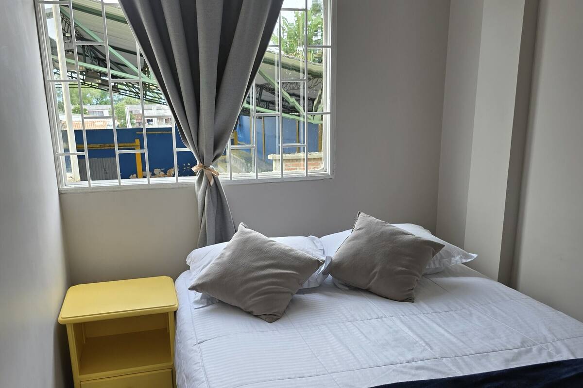 Un dormitorio con cama, mesita de noche, ventana con cortinas y vista a un edificio.