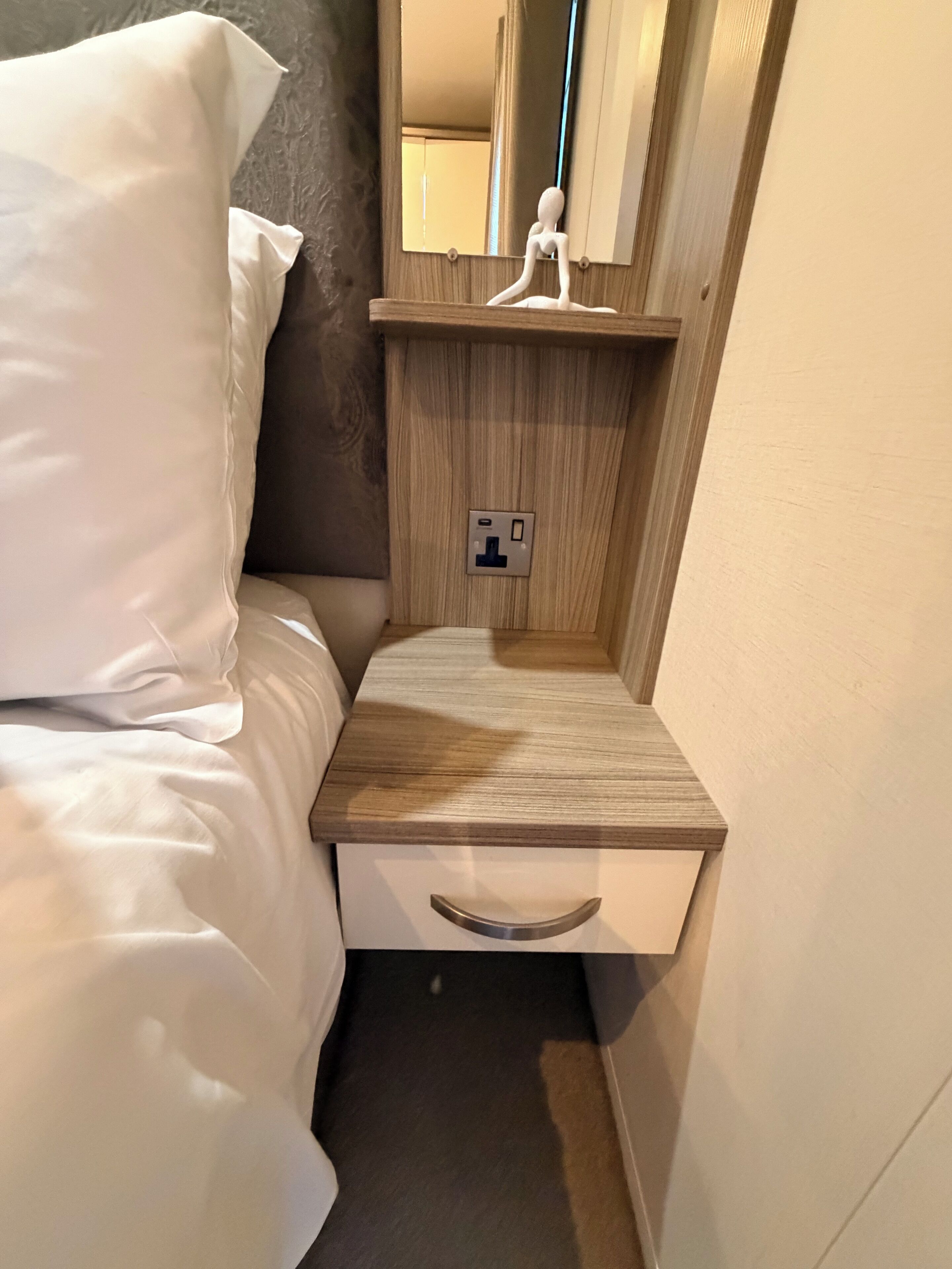 A compact hotel room with a bed, a bedside table with a drawer, a mirror, and a power outlet.