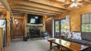 A wooden cabin interior with a fireplace, a television, a sofa, and a ceiling fan. - Cozy authentic log cabin with hot tub, Lake Access, near Sunday River & Mt Abram (Greenwood)
