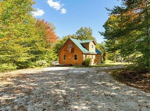 Une cabane en bois avec un toit vert, entourée d’arbres et bordée d’une allée de gravier.