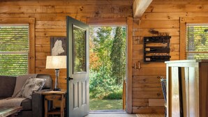 A wooden cabin interior with a sofa, a lamp, a door leading to a garden, and a shelf with glasses. - Cozy authentic log cabin with hot tub, Lake Access, near Sunday River & Mt Abram (Greenwood)