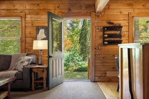 A wooden cabin interior with a sofa, a lamp, a door leading to a garden, and a shelf with glasses.