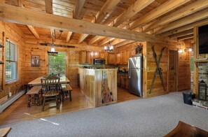 A log cabin interior with a wooden dining table, chairs, a kitchen with a refrigerator and microwave, and a fireplace.