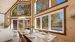 Interior de una cabaña de madera con una mesa de comedor para cuatro, ventanas grandes y vista a los árboles.
