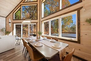 A wooden cabin interior with a dining table set for four, large windows, and a view of trees. - Cozy cabin w/ theatre, game room & hot tub (Sevierville)