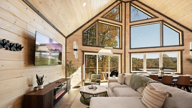 Un amplio espacio de sala con un sofá grande, una mesa de centro y un televisor montado en la pared. Varias ventanas ofrecen vistas al exterior.