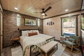 Une chambre à coucher aux murs lambrissés, comprenant un lit, un banc, une chaise, une petite table et un ventilateur de plafond.