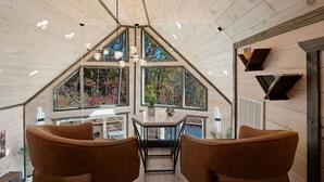 Una habitación con techo inclinado, ventanas amplias, una mesa con sillas y una lámpara colgante.