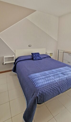 Ein Schlafzimmer mit einem Bett, einem Nachttisch, einer Lampe und einem kleinen Regal.