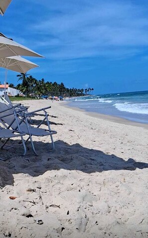Uma praia com cadeiras de praia e guarda-sóis, coqueiros e o oceano.