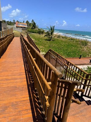 A wooden boardwalk leading to a beach with a view of the ocean and a building in the background. - Flat 003 | Studio Garden with pool views (Tamandaré)