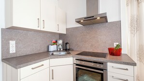 Una cucina compatta con mobili bianchi, un forno in acciaio inossidabile, un lavandino e un piano di lavoro con un bollitore e una ciotola rossa.