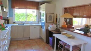 A kitchen with a dishwasher, a sink, a window with blinds, a table with chairs, and a trash bin. - Family house 5 minutes from the beach (Calella de Palafrugell)