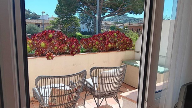 An outdoor patio with two wicker chairs and a table, a low wall with red flowering plants, and a view of buildings and trees in the background. - Family house 5 minutes from the beach (Calella de Palafrugell)
