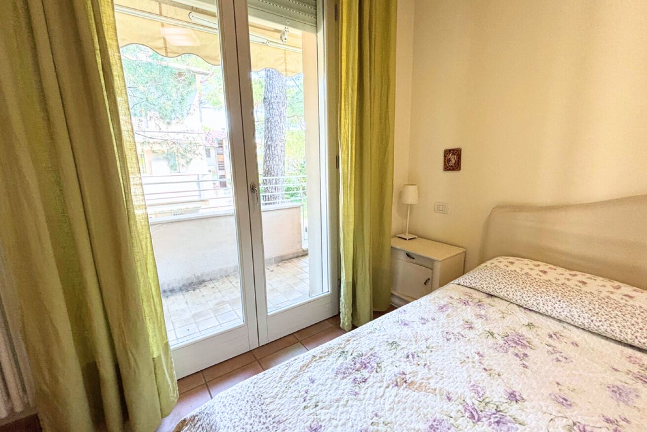 Quarto com cama, mesa de cabeceira, luminária, porta de vidro deslizante e vista para o exterior.