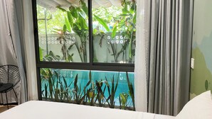 Una habitación con cama, ventana con vista a una piscina y plantas, y una silla.