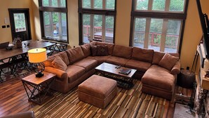 Un sofá modular grande en forma de L, una mesita y una lámpara en una habitación con ventanas amplias y una terraza de madera visible a través de las ventanas.