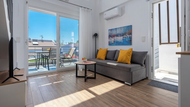A modern living room with a sofa, a small table, and a glass door leading to an outdoor area. - SLV Irida Apartment (Pangaio)